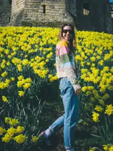 A bright, well-composed lifestyle portrait featuring a person standing in a field of daffodils with Warkworth Castle in the background. The image captures a clear, sunny day, effectively conveying a sense of seasonal travel and leisure.