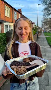 A creator reviews a large BBQ sharing tray from Big Norms, highlighting the variety of smoked meats including brisket, beef cheeks, turkey, and sausage, alongside sides like mac and cheese. The video features close-up shots of the food, the traditional smoker, and the creator enjoying the meal.