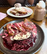 A high-quality, well-lit food photography shot featuring indulgent French toast dishes and a hot chocolate, likely taken at a brunch venue. The focus is sharp on the foreground plate, highlighting the textures of the toppings, while the background elements provide context without distracting from the main subject.