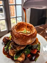 A high-quality, close-up action shot of a Sunday roast being served, featuring a gravy pour into a large Yorkshire pudding. The image is well-composed, using a 45-degree angle to showcase the layers of the meal, including roasted vegetables and meat. The lighting is bright and natural, highlighting the textures of the food, and the focus is sharp on the main subject.