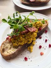 A close-up, top-down shot of a savoury brunch dish featuring scrambled eggs (likely tofu-based) on a slice of sourdough toast, topped with vegan bacon strips and pea shoots, served on a white plate.