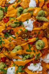 A high-quality, close-up food photography shot of a plate of loaded nachos. The image focuses on the textures of the melted cheese sauce, guacamole, sour cream, and jalapeños, making the dish look vibrant and appealing.