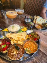 A high-angle, overhead shot of two Indian thali platters accompanied by cocktails, showcasing a variety of curries, rice, and naan bread in a restaurant setting.