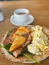 A close-up, top-down shot of a breakfast dish featuring a flatbread topped with spinach, bacon, scrambled eggs, and hash browns, served with a cup of coffee in the background.