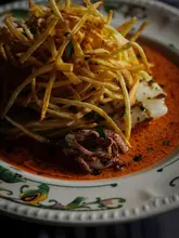 A high-quality, moody food photograph featuring a dish of squid topped with shoestring fries in a vibrant red sauce, served on an ornate, patterned plate. The lighting is dramatic and focused, highlighting the textures of the food while keeping the background dark and atmospheric, typical of high-end restaurant photography.