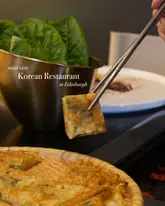 A close-up shot of a Korean pancake being held with chopsticks, with a blurred background showing a Korean BBQ setup.