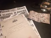A moody, atmospheric shot capturing the immersive Sherlock Holmes themed tasting menu experience at Six by Nico. The image features props from the interactive murder mystery element of the meal, including a case file and a themed newspaper, set against a dark table background.