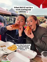 A casual, influencer-style selfie featuring two people eating hot dogs at an outdoor street food market. The image is cluttered with takeaway containers, detracting from the food.