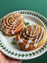A bright, overhead shot of two freshly baked cinnamon swirls drizzled with white icing, served on a vintage-style floral plate against a solid green background.