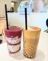 A bright, clean, and appealing shot of two drinks—a layered smoothie and an iced mocha—set against a blurred cafe background. The lighting is natural and flattering, highlighting the textures of the drinks.