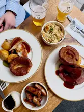 A high-quality, overhead flat-lay shot of a Sunday roast spread at a pub, featuring two main plates (beef and chicken), a side of cauliflower cheese, a side of pigs in blankets, and two glasses of beer. The lighting is natural and bright, highlighting the textures of the food.