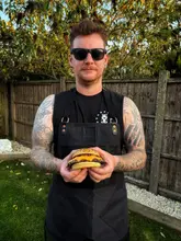 A portrait of a man wearing an apron, holding a homemade cheeseburger in a garden setting during golden hour.