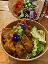 A top-down shot of street food items, specifically a chicken katsu curry bowl and a souvlaki pitta, served in takeaway packaging on a wooden table.