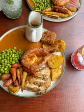 A high-angle, well-lit photograph of a Sunday roast dinner served in a pub setting. The plate features roast chicken, golden roast potatoes, a Yorkshire pudding, peas, carrots, and stuffing, accompanied by a gravy boat and a cocktail.