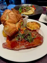 A close-up, slightly high-angle shot of a Sunday roast dinner served in a restaurant setting, featuring a large Yorkshire pudding, roast beef, and a side of macaroni cheese.