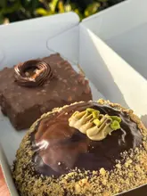 A high-quality, close-up shot of two artisan pastries—a Ferrero Rocher brownie and a pistachio-filled Greek brioche—nestled in a takeaway box. The natural lighting accentuates the glossy glaze and textures of the baked goods.