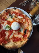 A top-down, overhead shot of a wood-fired pizza topped with a whole burrata ball, served on a dark wooden table with various condiments and cutlery surrounding the plate.