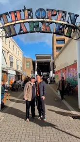 The video features two creators touring Brighton's Open Market, visiting multiple independent food and drink vendors. They sample a variety of items including Korean-Japanese fusion, sausage rolls, bagels, Greek food, and a self-built charcuterie board, highlighting the market's diverse and affordable offerings.