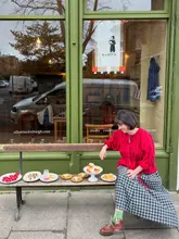 A lifestyle-focused shot featuring a person sitting on a wooden bench outside a cafe, with a selection of various pastries and tarts arranged on the bench in front of them.