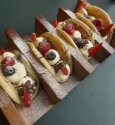 A high-quality, overhead shot of Nutella-filled taco pancakes served in a wooden rack, garnished with fresh strawberries, raspberries, and blueberries, dusted with icing sugar.