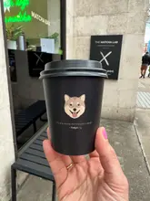 A close-up, eye-level shot of a branded black cup held in front of a cafe storefront, featuring a dog illustration and text.