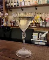 A close-up, eye-level shot of a Gibson cocktail served in a stemmed glass, placed on a wooden bar top with a blurred background of a bar shelf filled with various bottles.
