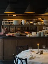 A moody, atmospheric interior shot of a steakhouse, featuring a neatly set dining table in the foreground with a candle, and an open kitchen grill area in the background displaying raw cuts of meat.