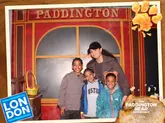 A family photo taken at The Paddington Bear Experience, featuring a branded frame overlay with the venue name and a graphic of a marmalade sandwich. The subjects are posing in front of a themed backdrop.