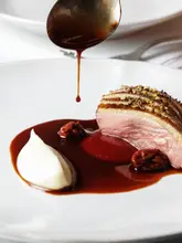 A professional, high-end food photography shot capturing a seared duck breast being drizzled with a rich, dark sauce, accompanied by a smooth puree and fruit garnish on a minimalist white plate.