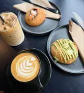 A clean, top-down flat lay photograph of a cafe table featuring two cookies, an iced coffee, and a hot latte with latte art. The image uses natural lighting and a dark, minimalist surface to make the textures of the food and drink stand out.