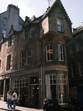 A high-quality, well-composed exterior shot of 'The Milkman' coffee shop in Edinburgh's Old Town, captured during a sunny day. The image highlights the charming, historic architecture of the venue and shows customers enjoying the outdoor seating area, creating an inviting atmosphere for a coffee break.