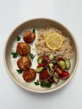 A clean, top-down, studio-style photograph of a homemade meal featuring Greek-style turkey meatballs, a generous dollop of tzatziki, a fresh tomato and cucumber salad, and a portion of rice. The lighting is bright and even, highlighting the textures of the food, and the presentation is simple and appetizing.