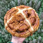 A close-up, high-quality shot of a hybrid pastry—a cinnamon roll topped with a hot cross bun cross, Biscoff sauce, and crushed biscuit crumbs—held in a hand against a blurred outdoor background.