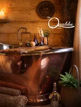 A high-quality, atmospheric shot of a copper bathtub inside a rustic cabin, styled with candles and towels to evoke a sense of luxury and relaxation.