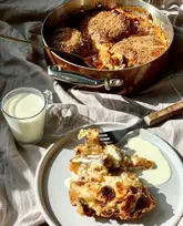 A top-down, natural light shot of a hot cross bun and butter pudding served in a copper pan, with a portion plated in the foreground alongside a glass of cream.