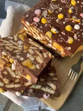 A high-quality, close-up shot of a homemade rocky road dessert topped with mini eggs. The lighting is natural and soft, highlighting the texture of the chocolate and biscuit chunks. The composition is effective, using a 45-degree angle to showcase the layers and the cross-section of the slice.