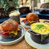 A close-up, eye-level shot of two smash burgers and a cast-iron pot of mac and cheese served on a wooden table.
