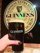 A close-up shot of a hand holding a pint of Guinness in front of a branded pub sign, likely taken in a dimly lit bar environment.