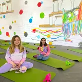 A photo of two children sitting on green yoga mats in an immersive art gallery space featuring a large, colourful wall mural of 'The Colour Monster'.