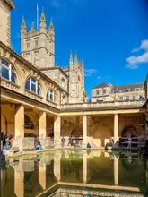 A high-quality, well-composed photograph of the Roman Baths in Bath, capturing the historic architecture and the pool under a clear blue sky. The image effectively showcases the venue's atmosphere and historical significance, making it an appealing destination for tourists and families.