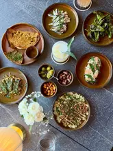 A high-quality, overhead flat-lay photograph showcasing a variety of small plates and a cocktail on a dark, textured table. The arrangement is balanced and visually appealing, highlighting the textures and colors of the food.