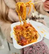 A close-up, first-person perspective shot of a fork lifting noodles from a takeaway container at a food festival. The focus is sharp on the food, with the creator's arm and a blurred background providing context.