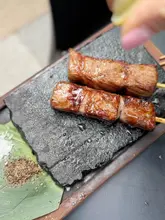 A high-quality, close-up shot of Wagyu beef skewers being finished with a squeeze of fresh lemon. The image is presented on a rustic slate board, effectively highlighting the texture and glaze of the meat. The shallow depth of field keeps the focus entirely on the food, creating an appealing and professional aesthetic.