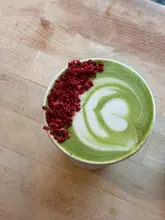 A top-down, close-up shot of a matcha latte in a takeaway cup, garnished with freeze-dried raspberries, set against a light wooden surface.