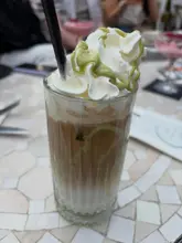 A close-up shot of a pistachio iced latte served in a tall glass with whipped cream and green drizzle, captured in a bright, airy orangery setting.