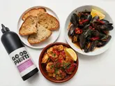 A high-quality, well-lit flat lay photograph featuring a bottle of 'Good Phats' Spanish extra virgin olive oil alongside a meal of gambas pil pil, harissa mussels, and crusty sourdough bread. The composition is clean and professional, effectively showcasing the product in a culinary context.