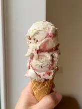 A high-quality, close-up shot of a double-scoop rhubarb crumble ice cream cone held in a hand. The lighting is soft and natural, effectively highlighting the creamy texture and swirls of the ice cream against a neutral, clean background.