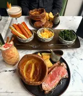 A high-quality, overhead shot of a Sunday roast spread at a restaurant, featuring two plates of roast beef with large Yorkshire puddings, roast potatoes, and gravy, accompanied by side dishes of roasted carrots and parsnips, cauliflower cheese, and kale, alongside two cocktails.