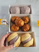A bright, top-down photograph showcasing three boxes of pastries from a bakery, including butter rice cakes and taro paste fluffy buns. A hand is visible in the frame, holding one of the buns to display its texture.