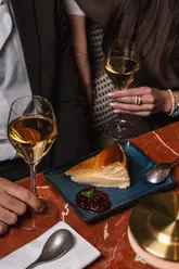 A high-quality, lifestyle-focused shot featuring a slice of Basque cheesecake with berry compote, served on a marble table alongside two glasses of white wine, capturing an intimate dining experience.